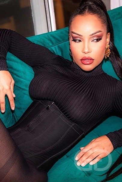 Beatriz, elite Latin escort, lying on a teal sofa in a black ribbed top and gold earrings